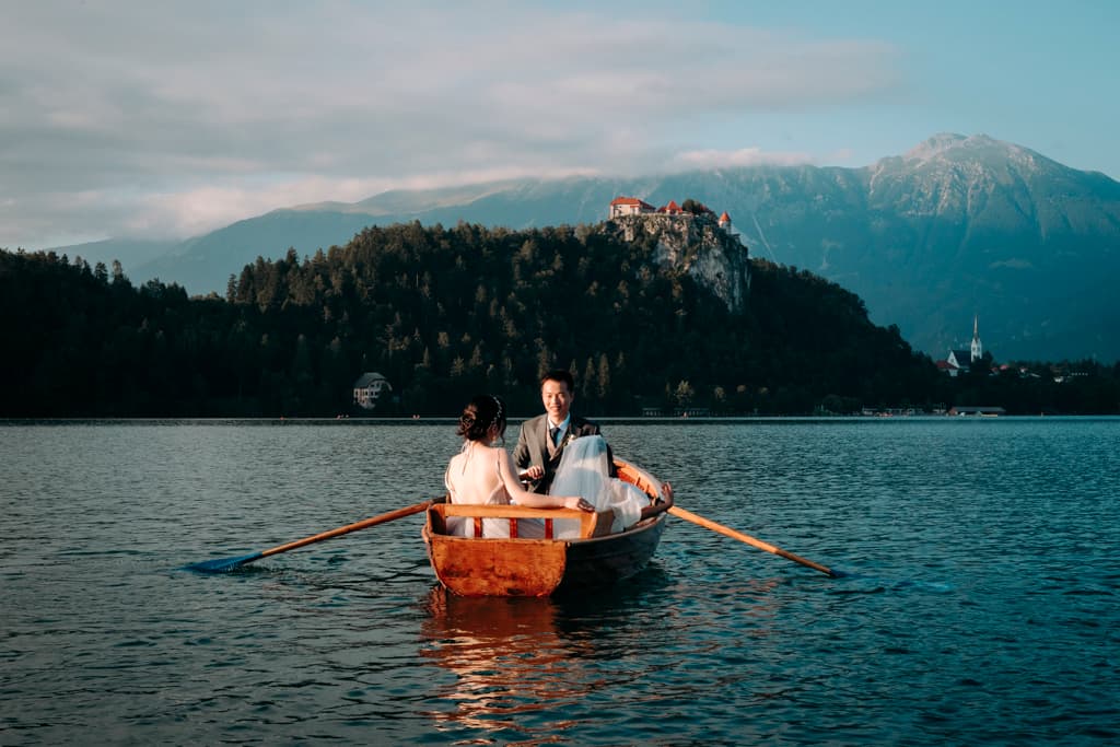 bride and groom on a boat on lake Bled with mountains and Church of the Assumption of the Blessed Virgin Mary in the background